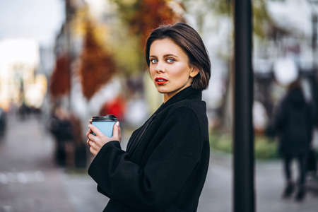 Young woman with short haircut and red lips, holds a cup of coffee, strolling through the streets of the city. Around are people and cars.の写真素材
