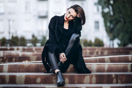 Full length portrait of young woman with short haircut and red lips, dressed in black. Woman sitting on the stairs outdoors on the stairsの写真素材