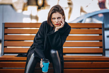 Young woman with short haircut and red lips, dressed in black coat, sits on a bench in the city and holds a cup of coffee.の写真素材