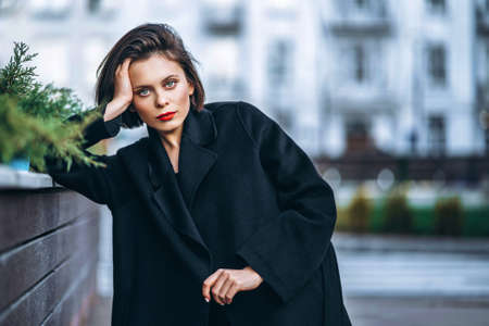 Close up portrait of young woman with short haircut and red lips, dressed in black. Woman posing, leaning on a wooden fence with plants. Blurred urban background.の写真素材