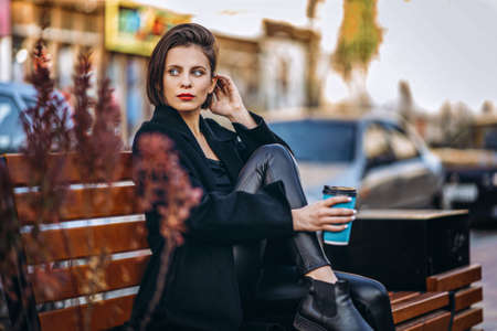 Young woman with short haircut and red lips, dressed in black coat, sits on a bench in the city and holds a cup of coffee.の写真素材