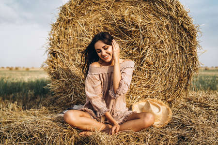 A young woman with long hair and in a dress sits near a hay bale. Woman posing smiling and looking at camera.の写真素材