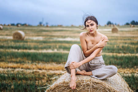 Brunette in linen pants and bare shoulders sitting on a hay bales in warm autumn day. Woman looking at camera. Behind her is a wheat field.の写真素材