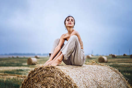 Brunette in linen pants and bare shoulders sitting on a hay bales in warm autumn day. Woman looking at camera. Behind her is a wheat field.の写真素材