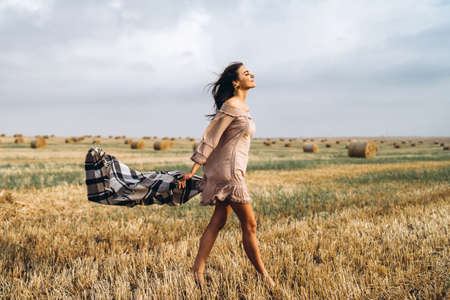 Full length portrait of a beautiful brunette in a dress and with a warm plaid. Woman enjoying a walk in a wheat field with hay bales.の写真素材
