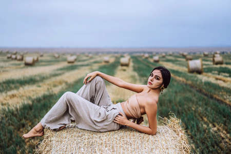 Young slender brunette girl with bright makeup in beige top with bare stomach and gray pants, lying on hay, posing for camera, photo shoot on field, blue skyの写真素材