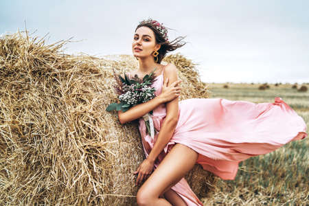 Young brunette in a pink satin dress, without linen standing near hay bales in windy weather. A woman holds a bouquet of wildflowers.の写真素材