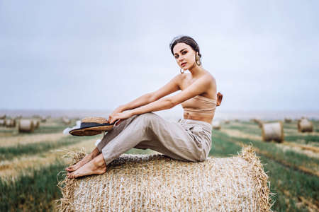 Barefoot brunette in linen pants and bare shoulders sitting on a hay bales in warm autumn day. Woman looking at camera. Behind her is a wheat field.の写真素材