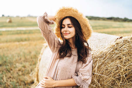 Closeup portrait of beautiful smiling woman with closed eyes. The brunette leaned on a bale of hay. A wheat field on the background.の写真素材