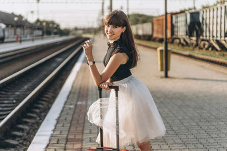 Female brunette traveler with red suitcase in white skirt waiting for a train and walking on raiway stationの写真素材