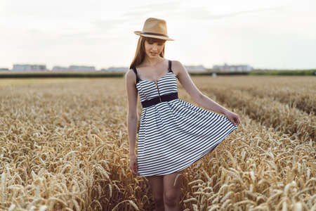 Pretty brunette girl in light dress wearing hat and walking outdoor in wheat field at sunset timeの写真素材