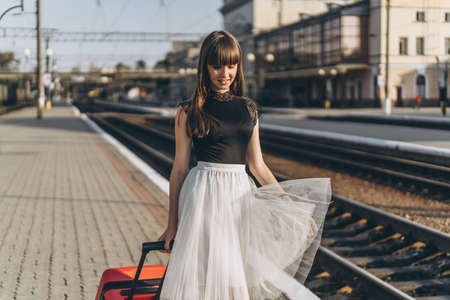Female brunette traveler with red suitcase in white skirt waiting for a train and walking on raiway stationの写真素材