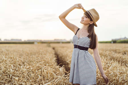 Pretty brunette girl in light dress wearing hat and walking outdoor in wheat field at sunset timeの写真素材