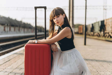 Female brunette traveler with red suitcase in white skirt waiting for a train on raiway stationの写真素材