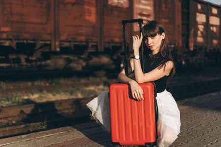 Female brunette traveler with red suitcase in white skirt waiting for a train on raiway station