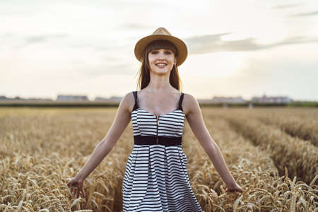 Pretty brunette girl in light dress wearing hat and walking outdoor in wheat field at sunset timeの写真素材