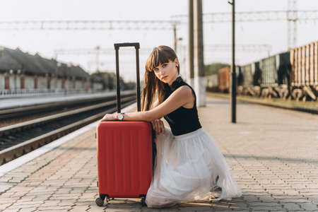 Female brunette traveler with red suitcase in white skirt waiting for a train on raiway stationの写真素材