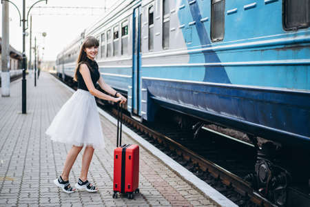 Female brunette traveler with red suitcase in white skirt waiting for a train and walking on raiway stationの写真素材