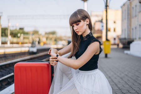 Female brunette traveler with red suitcase in white skirt waiting for a train on raiway station and checking time on her wristwatch