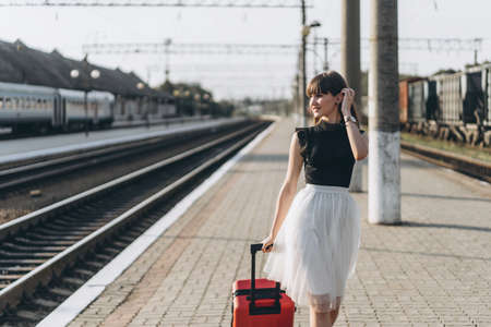 Female brunette traveler with red suitcase in white skirt waiting for a train and walking on raiway stationの写真素材