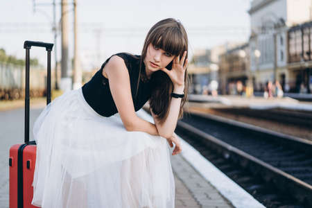 Female brunette traveler with red suitcase in white skirt waiting for a train on raiway stationの写真素材