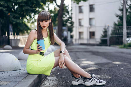 Pretty long hair brunette girl in green light dress with blue paper cup of beverage sitting outdoor on the street in the cityの写真素材
