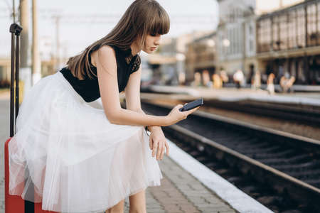 Female brunette traveler with red suitcase in white skirt waiting for a train on raiway station and using cellphoneの写真素材