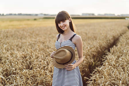 Pretty brunette girl in light dress wearing hat and walking outdoor in wheat field at sunset timeの写真素材