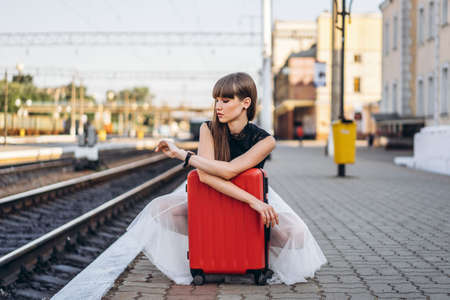 Female brunette traveler with red suitcase in white skirt waiting for a train on raiway station and checking time on her wristwatchの写真素材