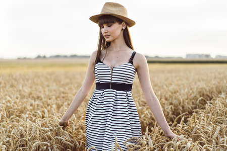 Pretty brunette girl in light dress wearing hat and walking outdoor in wheat field at sunset timeの写真素材