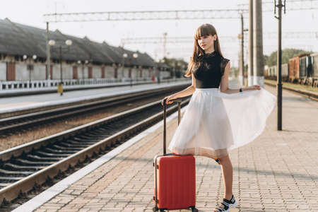 Female brunette traveler with red suitcase in white skirt waiting for a train and walking on raiway stationの写真素材
