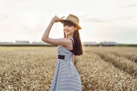 Pretty brunette girl in light dress wearing hat and walking outdoor in wheat field at sunset timeの写真素材