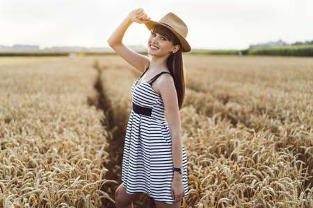 Pretty brunette girl in light dress wearing hat and walking outdoor in wheat field at sunset timeの写真素材
