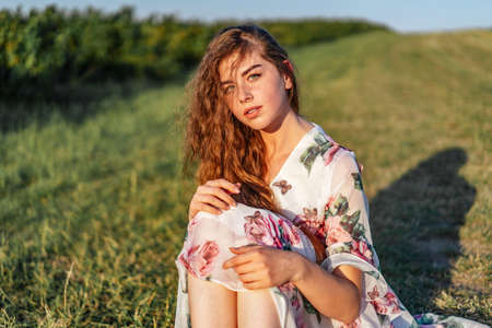 Young girl with long curly hair, brunette, face in freckles, with makeup and green eyes, in light white dress posing for camera, background blurred, natureの写真素材
