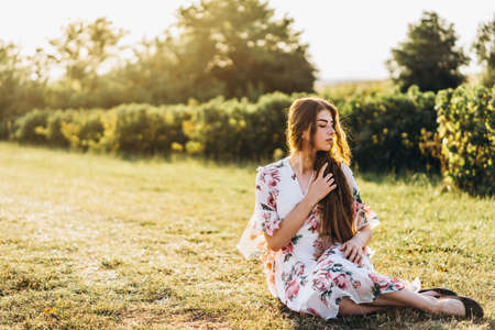 Full length portrait of beautiful woman with long curly hair on currant field background. Girl in a light dress sits on the grass in sunny day.の写真素材