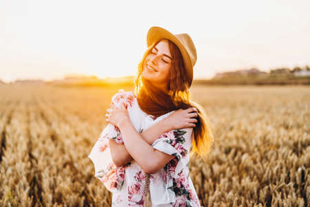 Incredible young woman with long curly hair and freckles face. Woman in dress posing in wheat field at sunset. Close up portrait.の写真素材