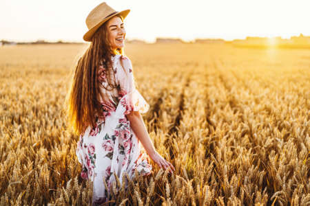 Portrait of a beautiful young woman with curly hair and freckles face. Woman in dress and hat posing in wheat field at sunset and looking at camera.の写真素材