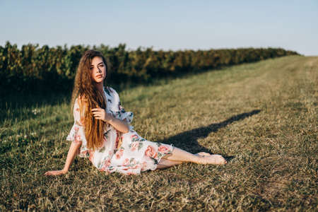 Full length portrait of beautiful woman with long curly hair on currant field background. Girl in a light dress sits on the grass in sunny day.の写真素材