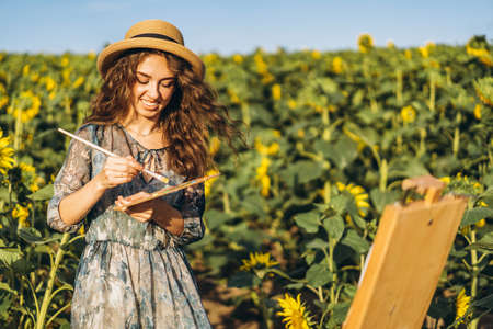 A young woman with curly hair and wearing a hat is painting in nature. A woman stands in a sunflower field on a beautiful day.の写真素材