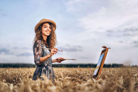 Portrait of smiling female artist with curly hair in hat. Girl draws a picture of a landscape in a wheat field. Copy space.の写真素材