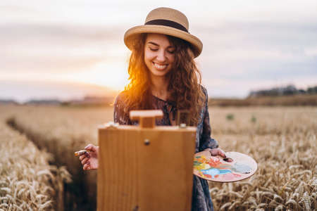 Beautiful girl artist paints in a wheat field. Woman smiles, holds a brush and palette in hands, in front of her stands an easel. Behind her is orange sunlight.の写真素材