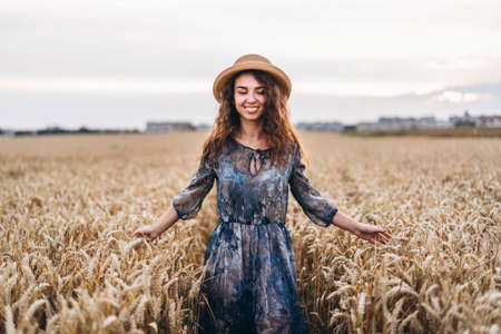 Closeup portrait of a beautiful young woman with curly hair. Woman in dress and hat standing in wheat field.の写真素材