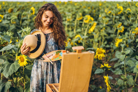 A young woman with curly hair and wearing a hat is painting in nature. A woman stands in a sunflower field on a beautiful day.の写真素材