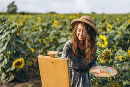 A young woman with curly hair and wearing a hat is painting in nature. A woman stands in a sunflower field on a beautiful day.の写真素材