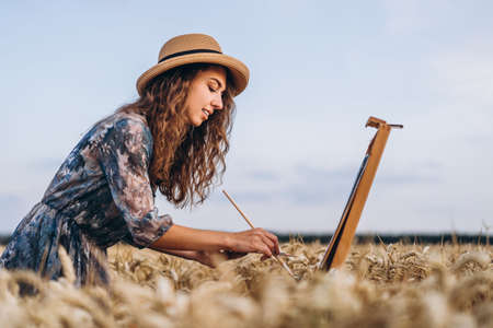 Portrait of smiling female artist with curly hair in hat. Girl draws a picture of a landscape in a wheat field. Copy space.の写真素材
