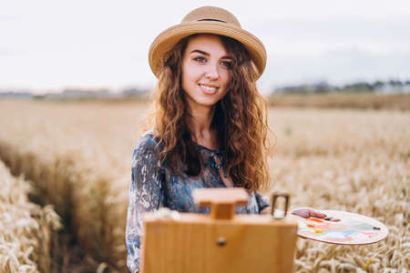 Portrait of smiling female artist with curly hair in hat. Girl draws a picture of a landscape in a wheat field.の写真素材