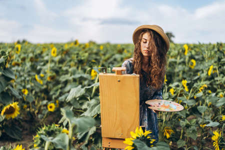 A young woman with curly hair and wearing a hat is painting in nature. A woman stands in a sunflower field on a beautiful day.の写真素材