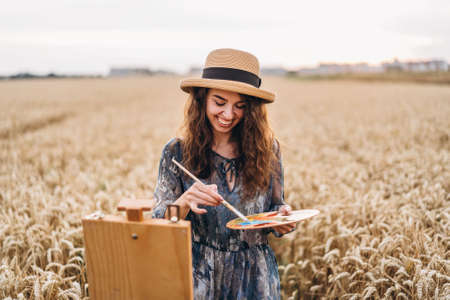 Portrait of smiling female artist with curly hair in hat. Girl draws a picture of a landscape in a wheat field. Copy space.の写真素材