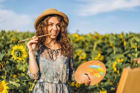A young woman with curly hair and wearing a hat is painting in nature. A woman stands in a sunflower field on a beautiful day.の写真素材