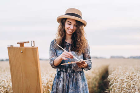 Portrait of smiling female artist with curly hair in hat. Girl draws a picture of a landscape in a wheat field. Copy space.の写真素材